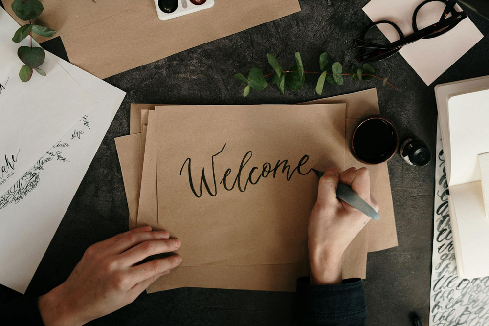 Hands writing the word "Welcome" on a piece of paper at a desk, symbolizing employee onboarding and the first 12 months.